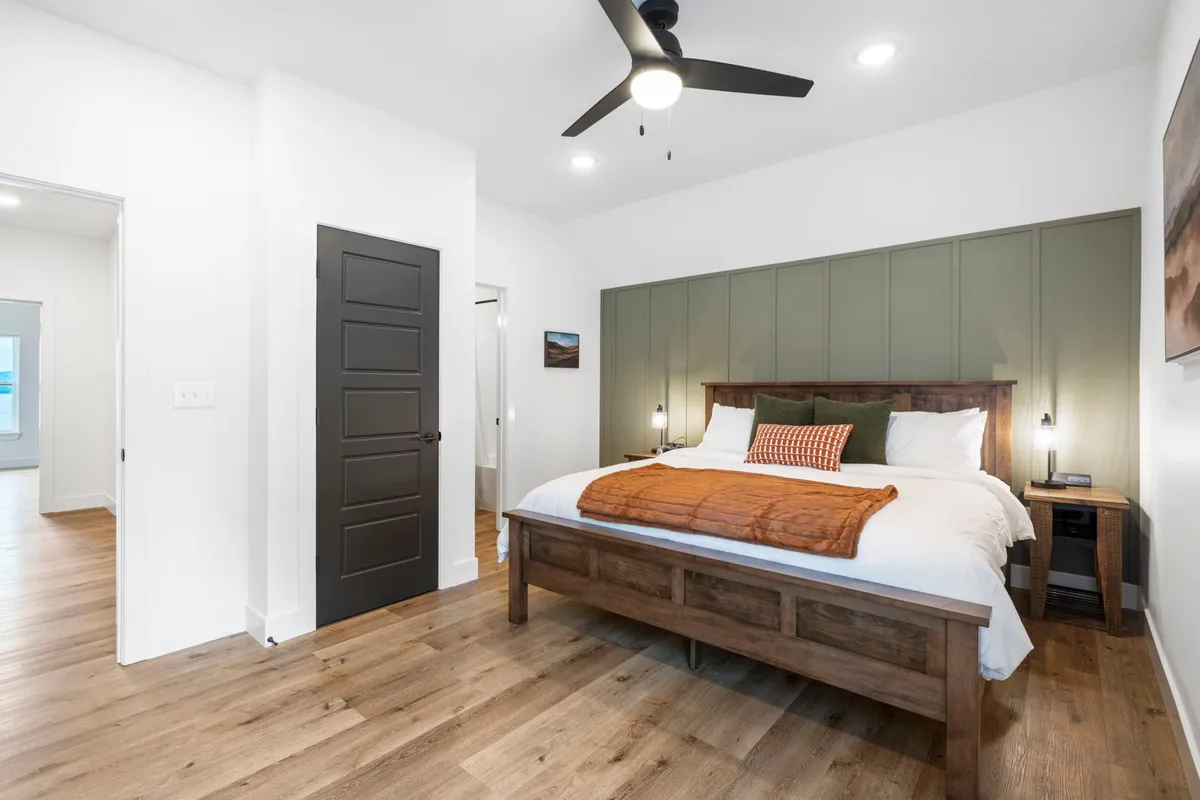 A serene bedroom featuring a bed with a wooden frame and a green headboard, accented by an orange throw blanket and nightstands with softly glowing lamps. Natural light filters into the space, creating a warm and inviting atmosphere perfect for relaxation.