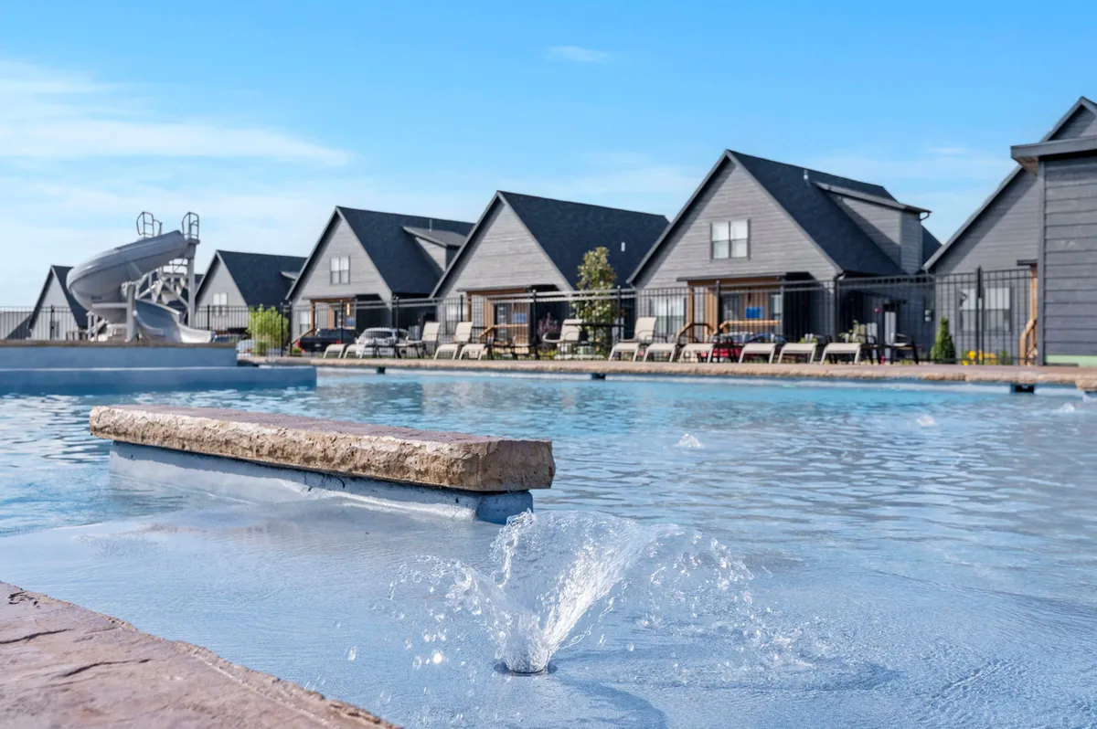 A vibrant exterior image of a resort-style pool with a water fountain and water slide, surrounded by modern homes. The pool area is lively, welcoming groups for swimming, sunbathing, and social gatherings.