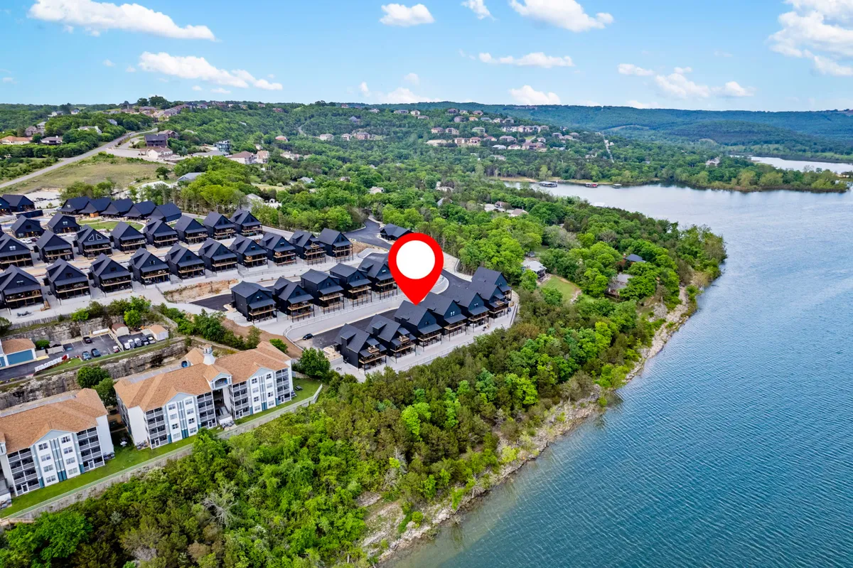 This image presents a tranquil shoreline along Table Rock Lake, with a row of modern black-roofed houses, lush green foliage, and the shimmering water stretching into the distance. The scene captures both the natural beauty of the lake and the inviting residential environment.
