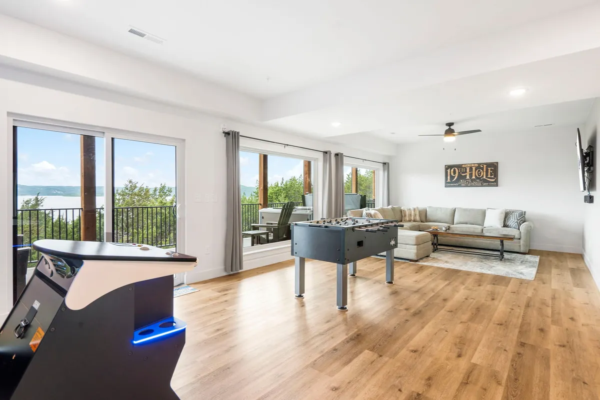 An energetic interior scene featuring a foosball table, comfortable sectional sofa, and large windows with views of the outdoors. The playful ambiance is enhanced by the presence of '19 Hole' wall art and a spacious balcony ideal for gathering.