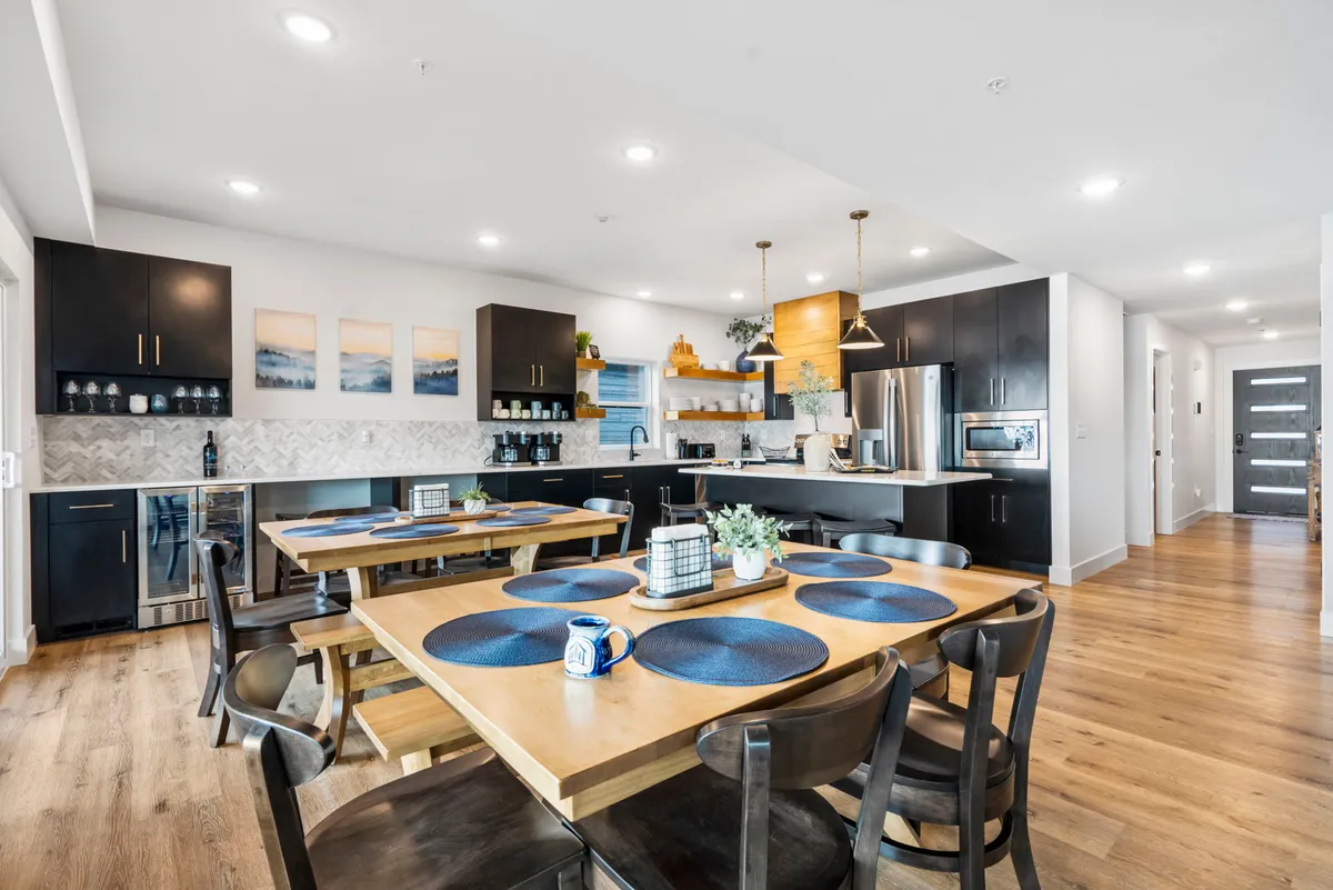 A view of the dining area featuring a large table perfect for group meals, with adjacent kitchen counter space and modern pendant lighting. Artwork and natural elements provide warmth, while the open-plan design connects dining and kitchen for seamless entertaining.