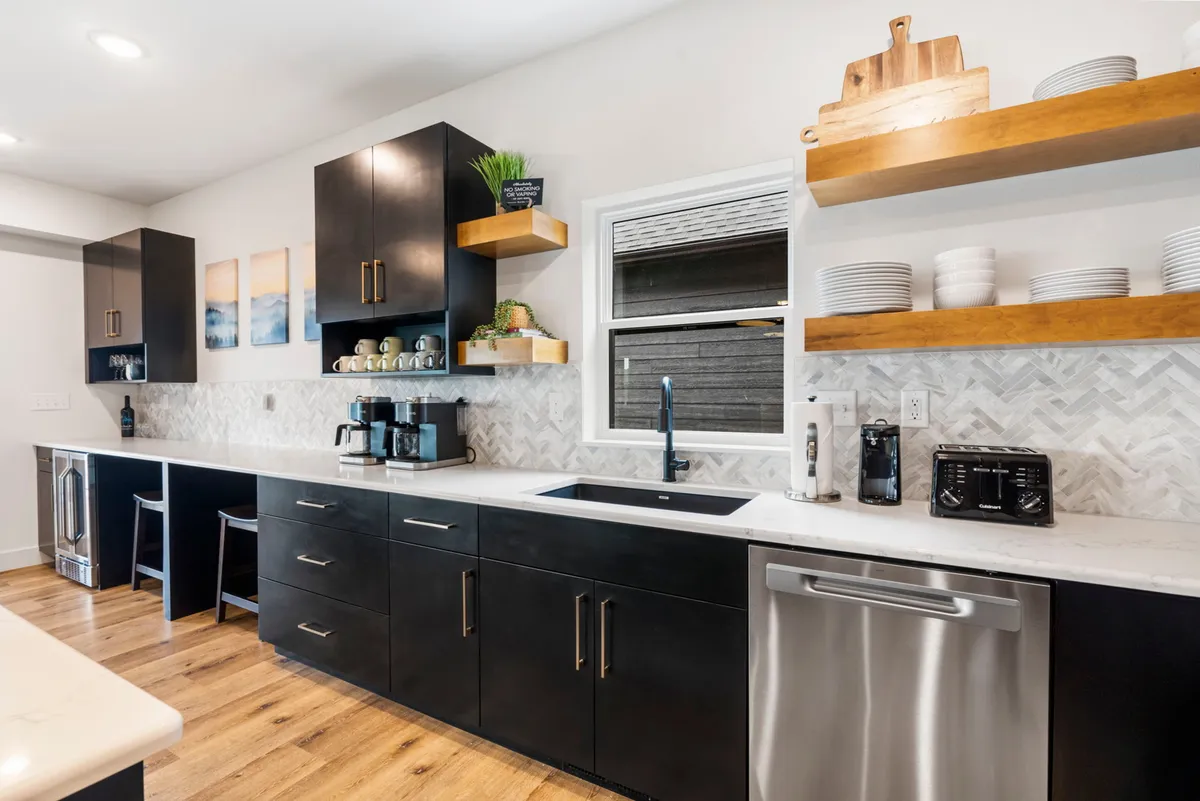 A stylish, contemporary kitchen with black cabinetry, white countertops, herringbone backsplash, and open shelves stocked with dishware. A dedicated coffee station and ample counter space hint at high-end appliances and culinary capabilities.