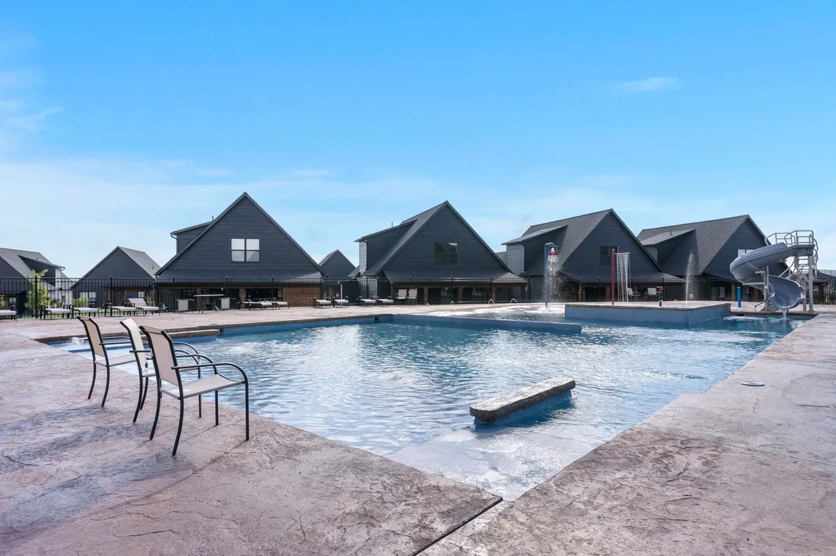 A lively community outdoor pool area with a water slide, splash pad, and sun loungers surrounded by buildings. The atmosphere is cheerful and energetic, inviting group fun, swimming, and warm-weather relaxation.