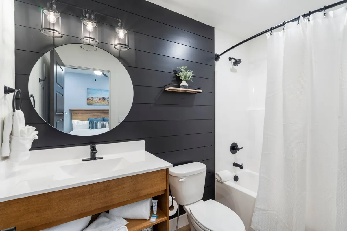 A contemporary bathroom with a circular mirror above a wooden vanity, a striking black accent wall, and modern light fixtures. The space feels serene and spa-like, ideal for refreshing morning routines or relaxing evening rituals.