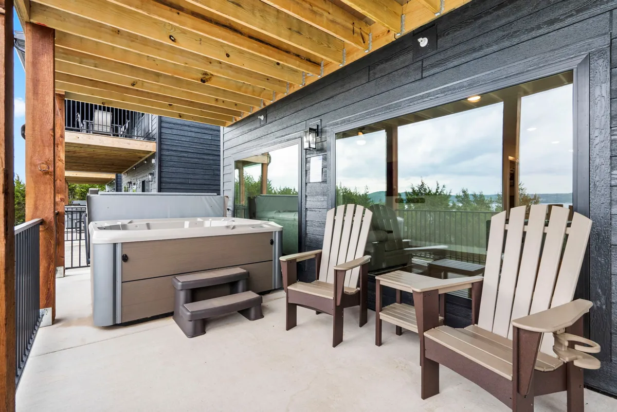 This exterior image showcases a private hot tub surrounded by Adirondack chairs, set on a patio with large windows opening to the natural landscape. The hot tub area overlooks the lake, creating an inviting space for relaxation and socializing while enjoying picturesque views.