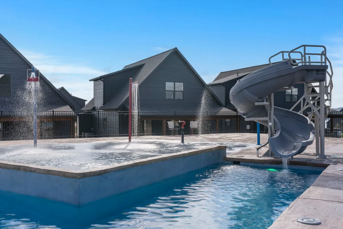 A vibrant exterior shot of a swimming pool with a water slide and splash pad, surrounded by community buildings. Children and families are visible enjoying playful moments, capturing the fun and active element of the resort's amenities.