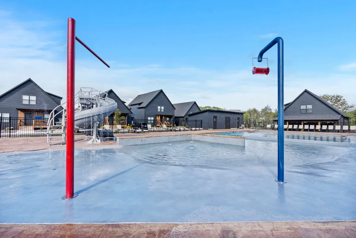 An exterior image showing a lively pool area with a water slide, spray features, and modern buildings in the background. Children and families are seen enjoying the water, suggesting fun, relaxation, and year-round recreation.